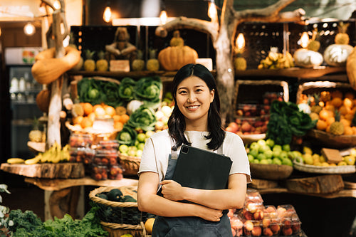 Smiling entrepreneur holding a digital tablet standing at an outdoor market. Asian woman in an apron looking at camera while standing against a street food market.