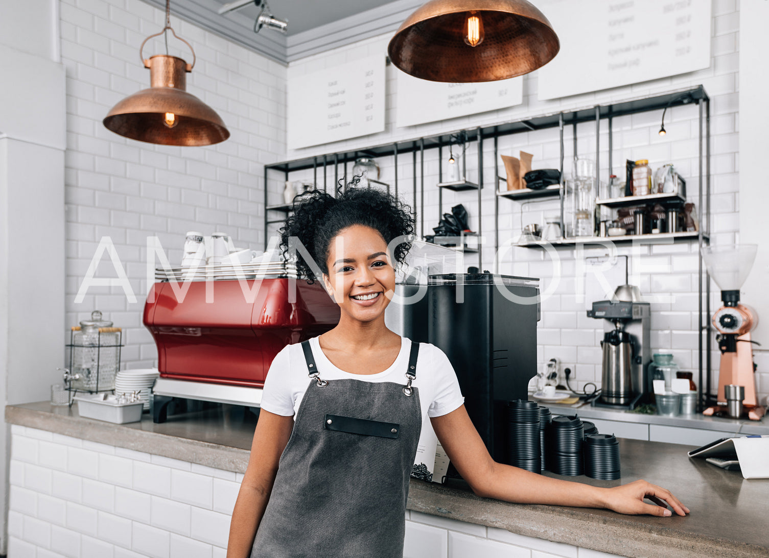 Young smiling woman with curly hair working as a barista. Female in an apron standing at a counter in a coffee shop and looking at camera.