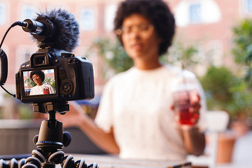 Woman with a juice in one hand talking to digital camera. Vlogger recording content.