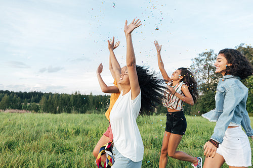 Group of multi-ethnic female friends raised hands up and throwing confetti while running on a field