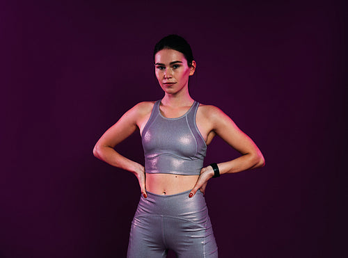 Smiling woman athlete in silver sportswear with hands on hips standing at magenta backdrop