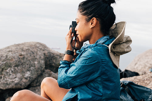 Side view of a woman taking photographs on her film camera while sitting on a rock during a mountain hike