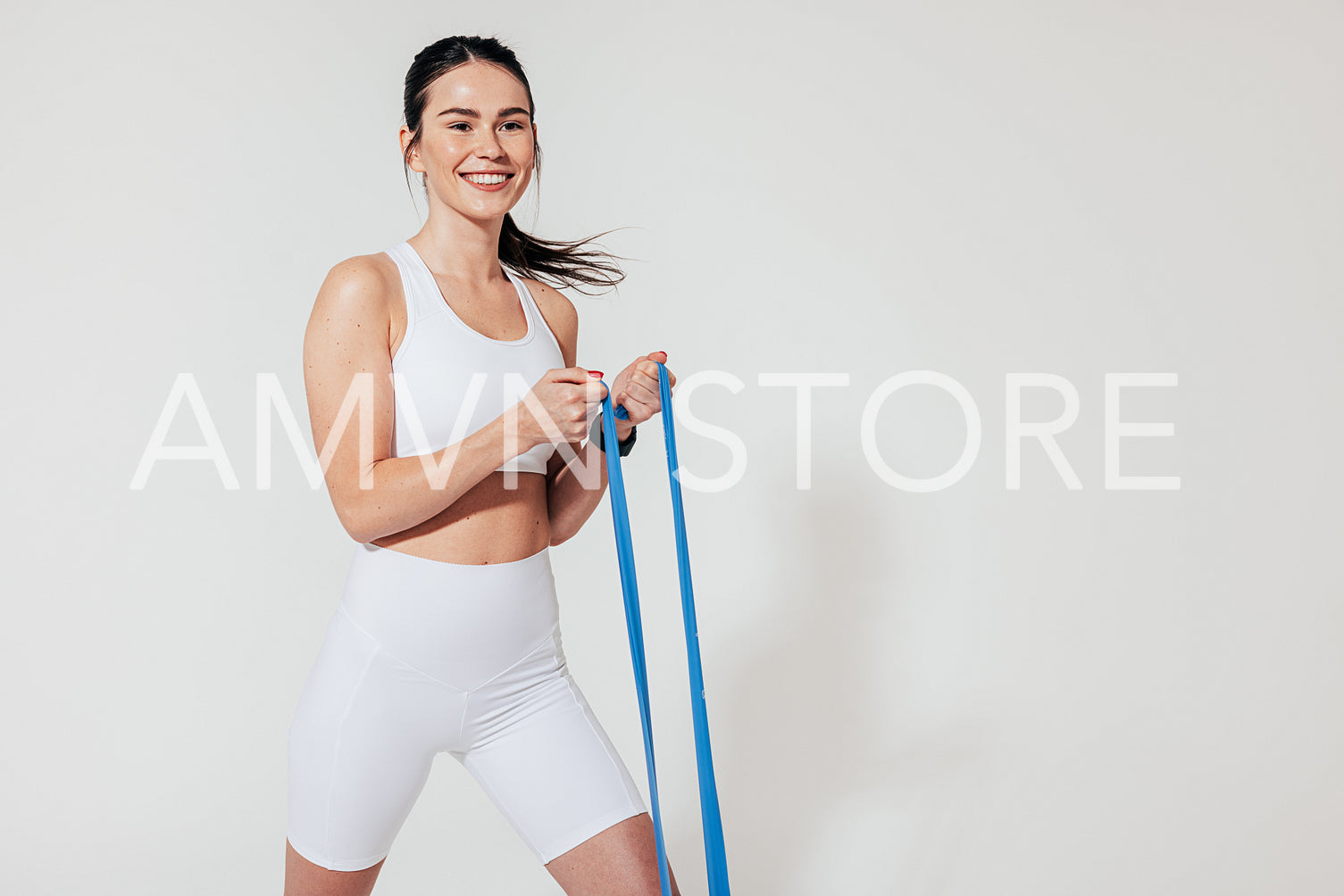 Cheerful female in white fitness attire exercising with blue resistance band at a white wall