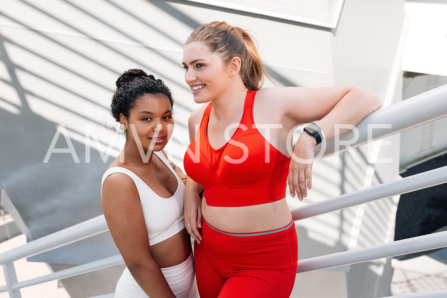 Two smiling females wearing fitnesswear standing on a bridge. Positive plus-size women relaxing during workout outdoors.