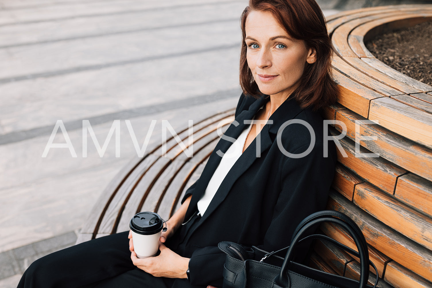 Middle-aged smiling businesswoman sitting on a bench outdoors. Female in black formal wear relaxing outdoors holding a coffee cup.