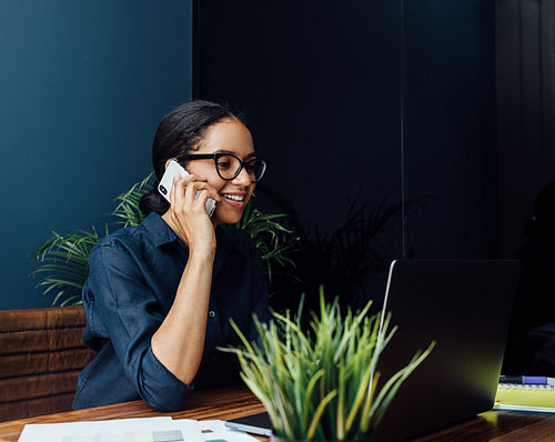 Smiling woman sitting at desk looking at laptop screen and talking over mobile phone