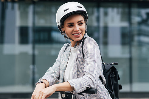 Young businesswoman with electric scooter looking away. Cheerful female in safety helmet wearing backpack standing outdoors.