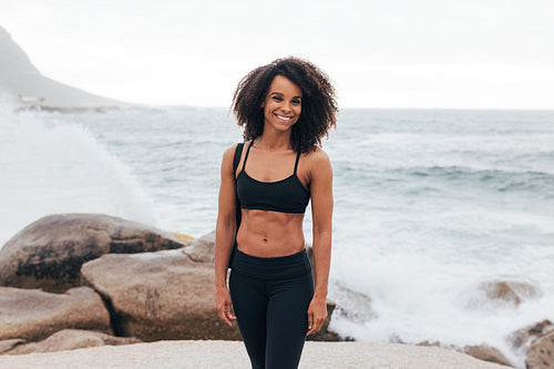 Smiling female relaxing after yoga. Muscular woman standing on rocks by ocean.