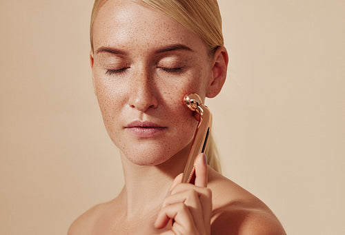 Young woman with closed eyes using face massager. Female with smooth freckled skin using face roller against a beige background.
