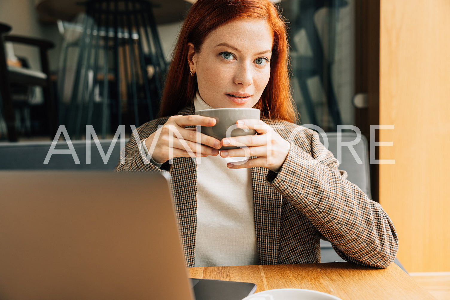 Beautiful woman with ginger hair looking at the camera in a cafe. Businesswoman in a blazer holding a cup sitting at a table.