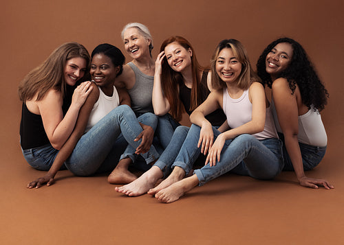 Six women of different ages sitting together in studio on brown background. Multi-ethnic group of diverse females having good times.