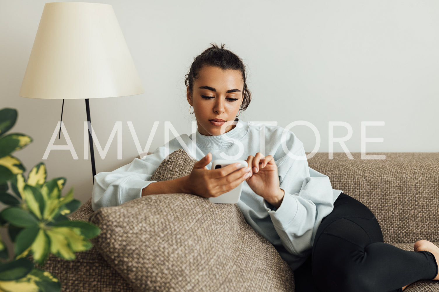 Young woman in casuals lying on a sofa in a living room holding a smartphone