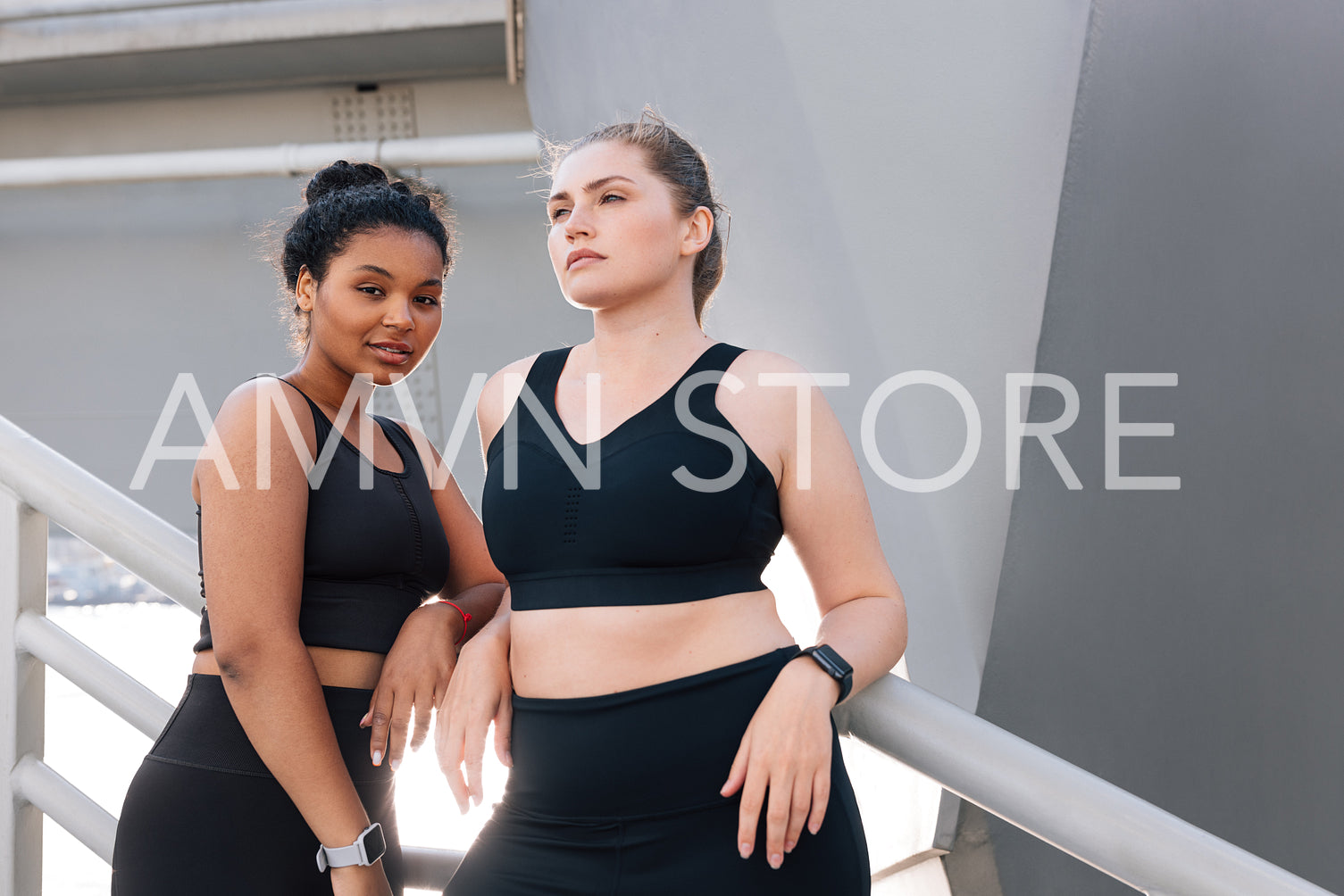 Two plus-size females in black fitness wear posing together after a workout. Young female in fitness attire looking at the camera while standing outdoors with her friend.