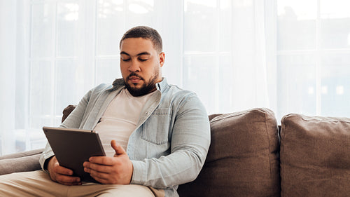 Young man in casual clothes sitting on sofa at home and using digital tablet