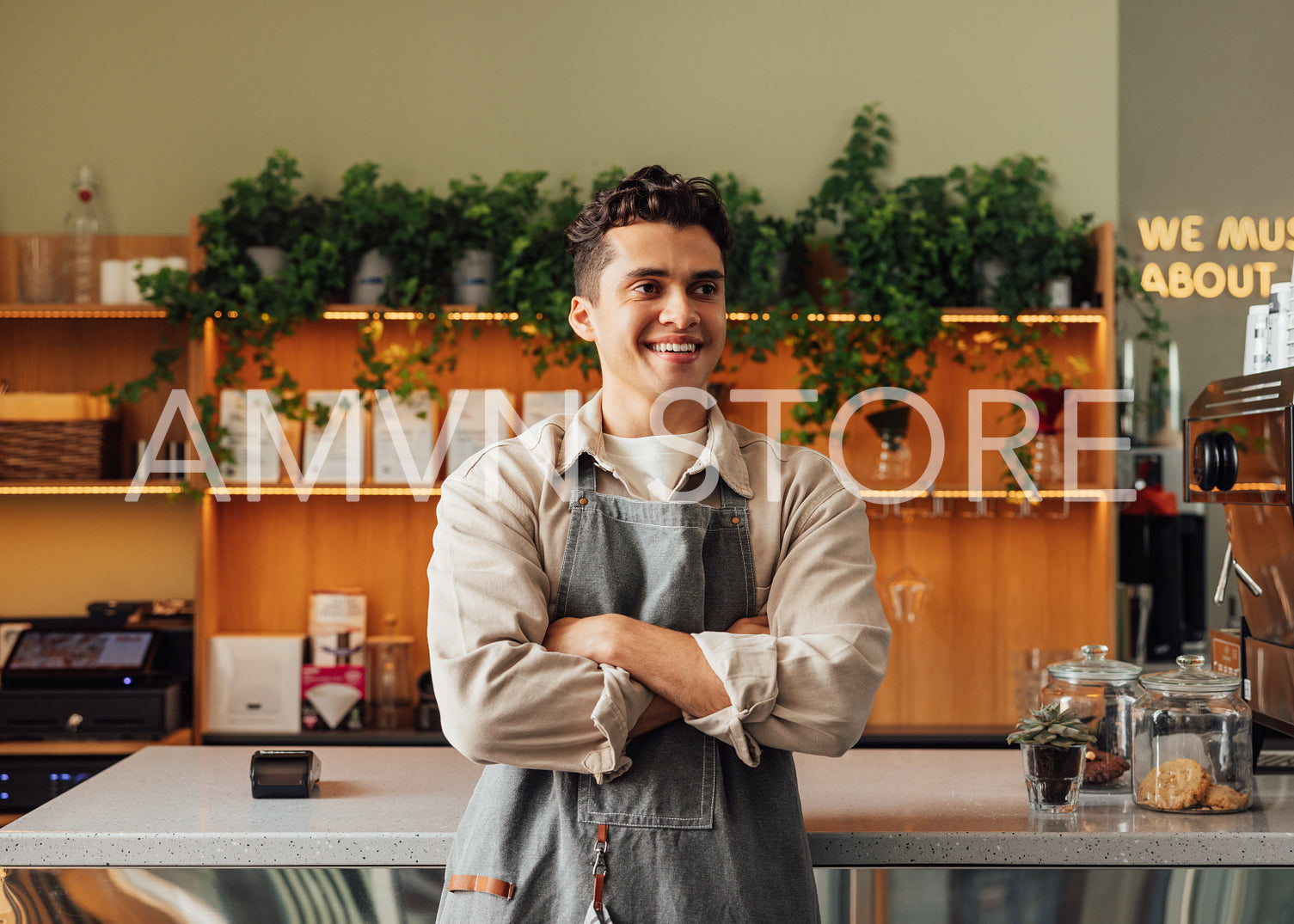 Confident barista in an apron. Young guy working as a barista in a coffee shop standing with crossed hands and looking away.