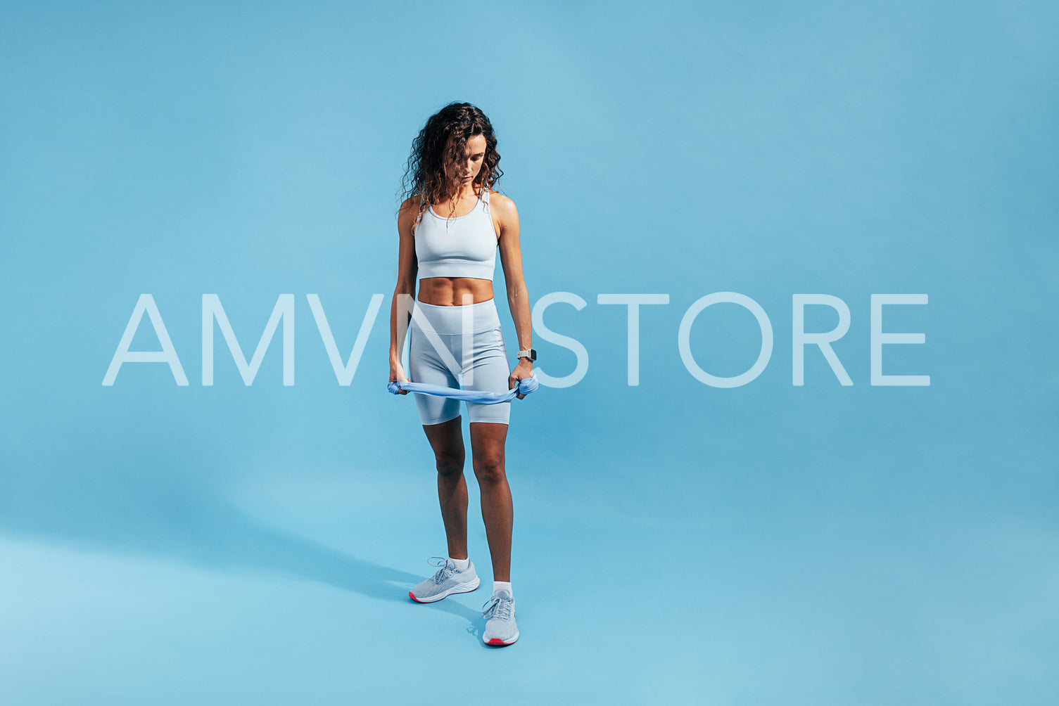 Young slim woman in fitness attire holding a resistance band and looking down on blue background