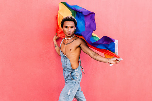 Young stylish man holding a waving LGBT flag against pink wall outdoors and looking at camera