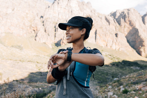 Smiling woman standing in valley holding smart watch on wrist. Female hiker in fitness wear exploring new path.