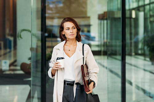 Middle-aged businesswoman holding coffee going out from the business building. Confident businesswoman with ginger hair at business building.