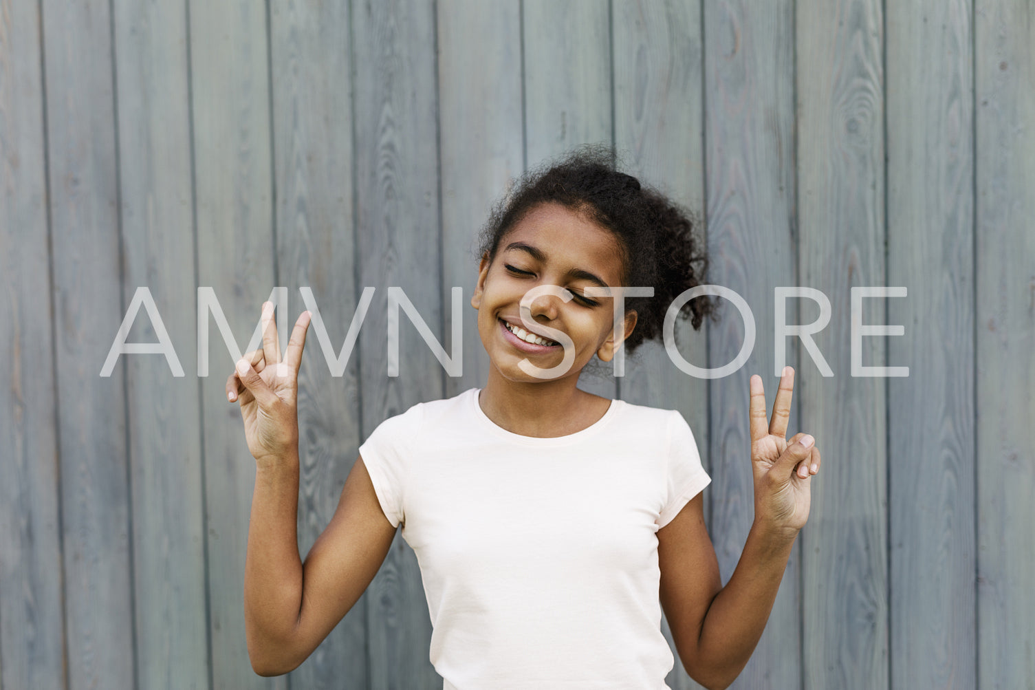 Cute girl showing victory sign, standing near a wall with eyes closed	