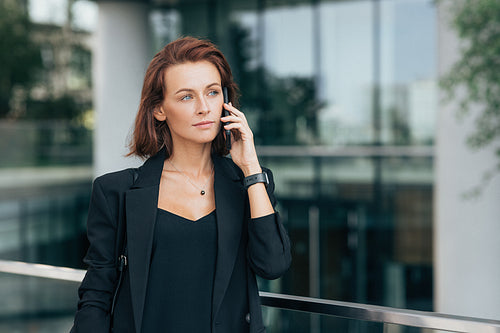 Confident businesswoman talking on mobile phone and looking away. Middle-aged female in black formal clothes making conversation on smartphone against glass building.