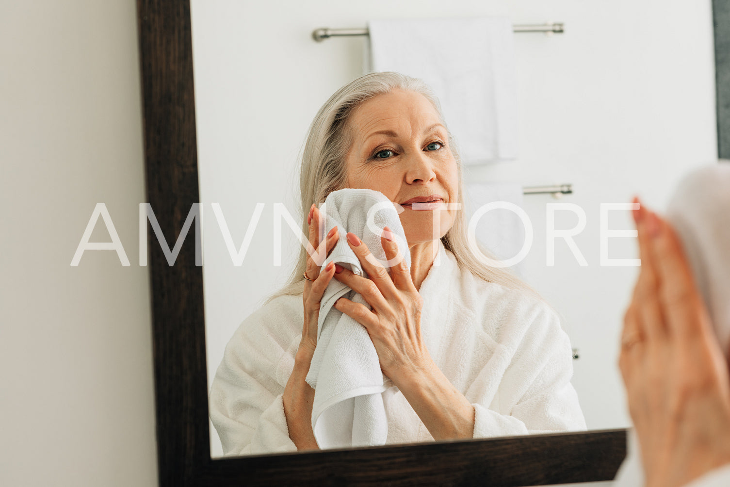 Senior woman with long hair wiping her face with a towel. Aged female in bathrobe doing morning routine in the bathroom.