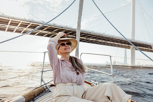 Mature woman in sunglasses puts on hat sitting on a sailboat at sunny day