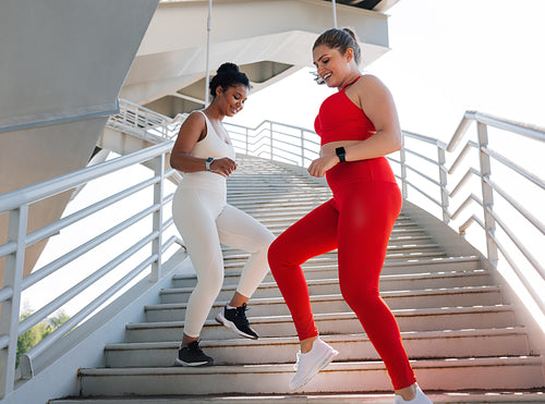 Two young plus-size females running together downstairs while practicing outdoors. Smiling women doing workouts together.