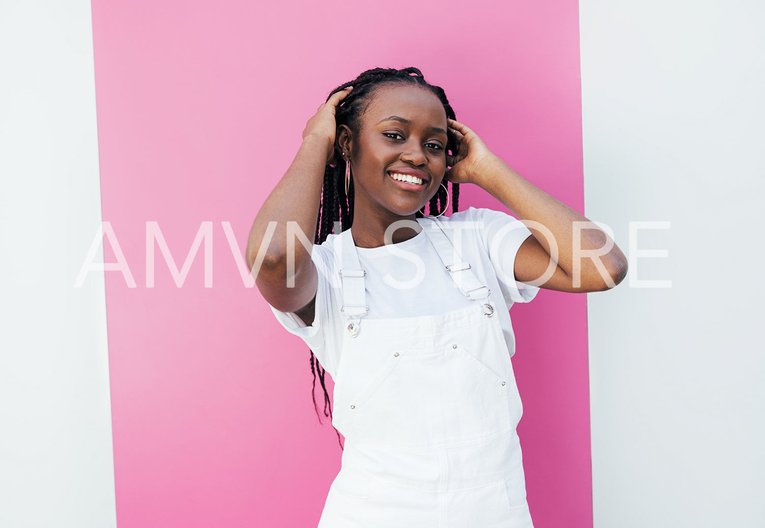 Smiling woman with braids adjusting hair while standing at white wall with pink stripe