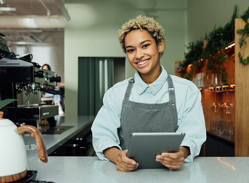 Smiling female barista with digital tablet in the cafe. Portrait of a young female in an apron at a counter in a coffee shop.