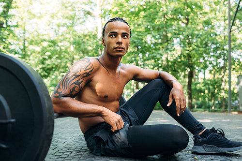 Muscular athlete sitting on sports ground with hand on barbell looking away