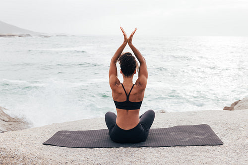 Young woman with hands raised practicing yoga on a beach at sunset. Back view of unrecognizable female meditating.