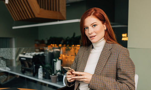 Portrait of a young businesswoman with ginger hair in a cafe. Female in formal wear holding a smartphone looking at camera.