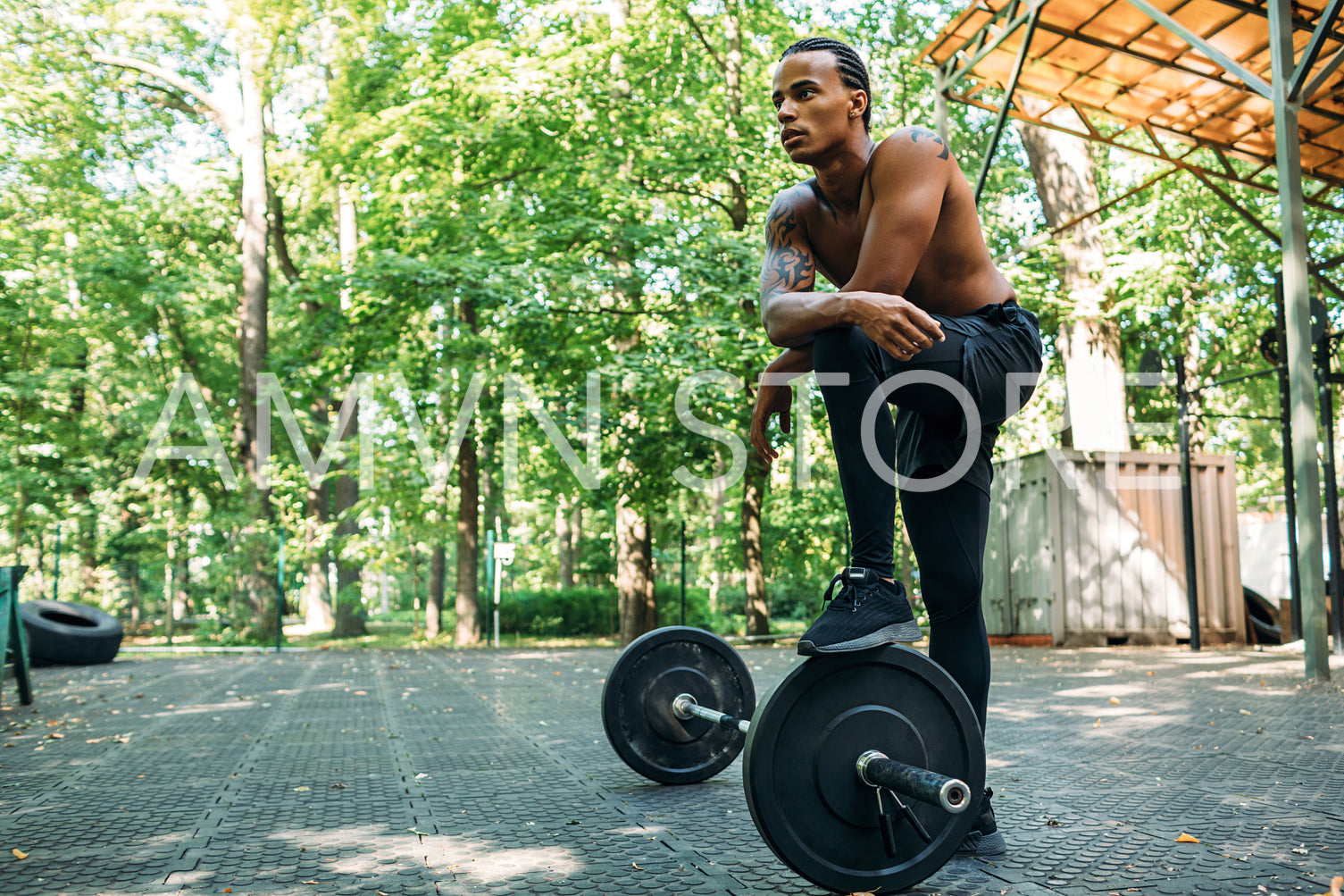 Bare-chest bodybuilder resting after deadlifting exercises at the sports ground. Young athlete stands with one leg on the barbell.	