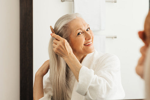 Cheerful senior woman combing her hair looking at a mirror in bathroom