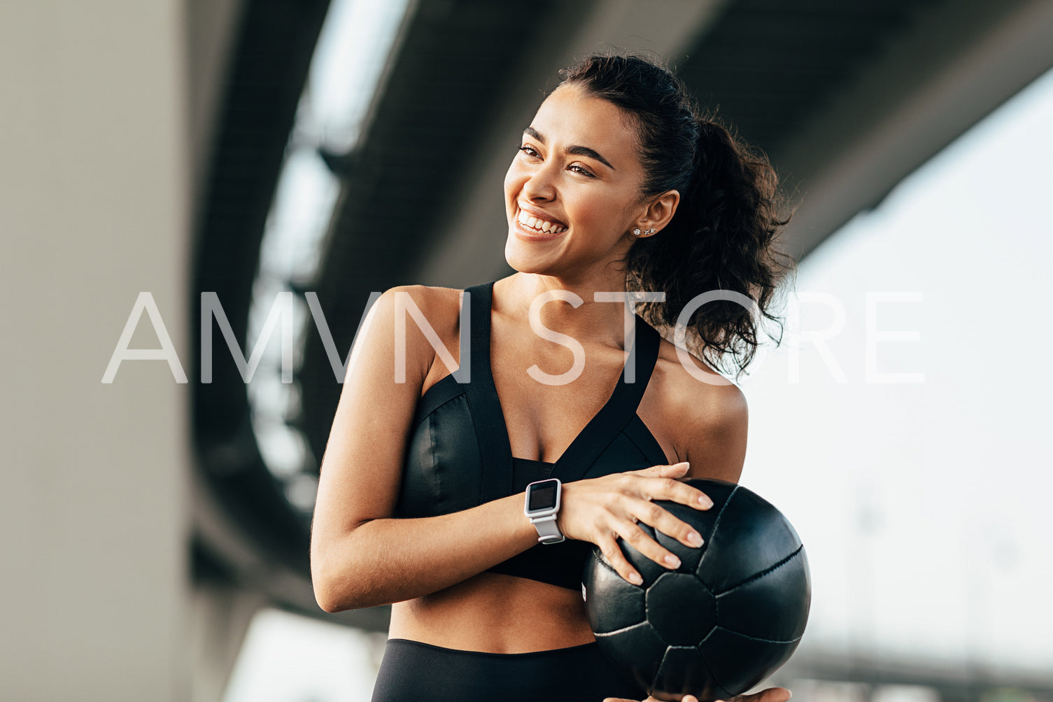 Beautiful woman holding a medicine ball and looking away. Smiling sportswoman exercising under a highway.	