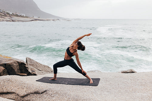 Young woman practicing Reverse Warrior pose by ocean at sunset. Female flexing her body on mat outdoors.