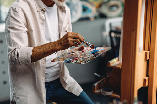 Male hand holding paintbrush and mixing oil paints on colorful messy palette