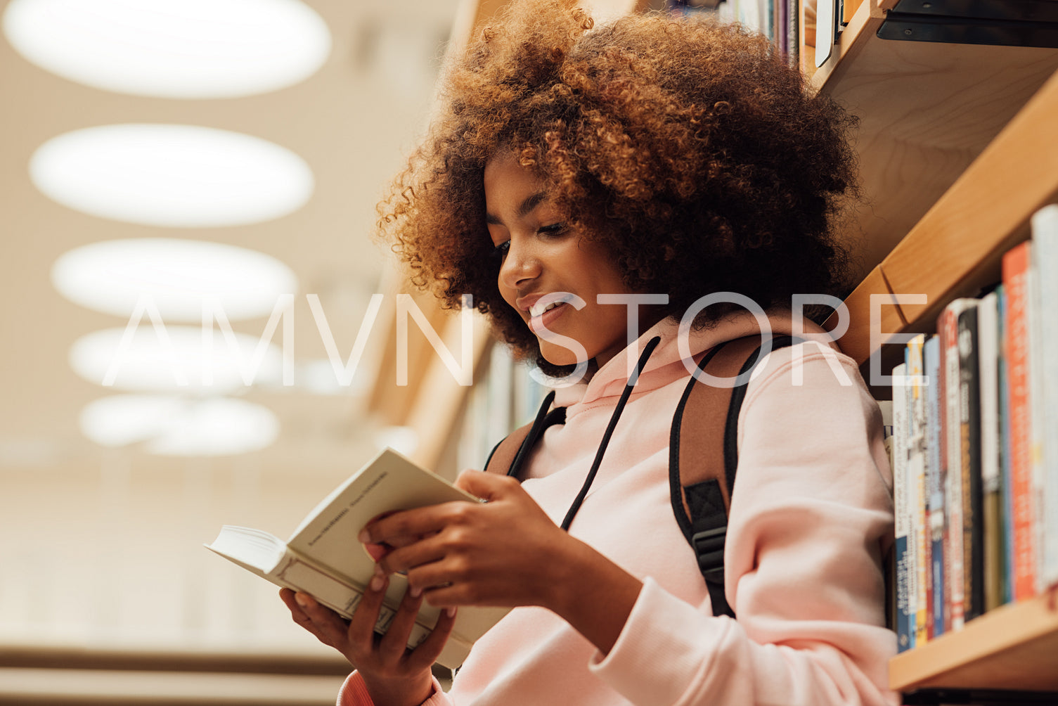 Girl in casuals with backpack leaning bookshelf in library. Student reading a book while standing in library.