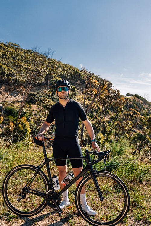 Male cyclist with bicycle in wild terrain looking at camera. Athlete in black sportswear with road bike relaxing.