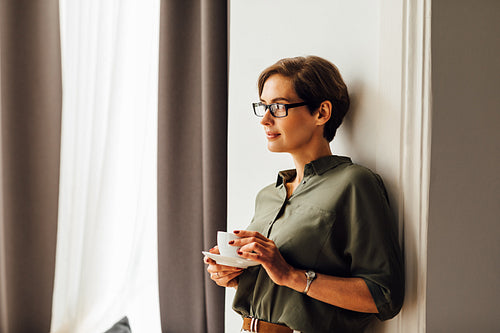 Mid adult caucasian woman in formal clothes standing in apartment room and having a cup of coffee