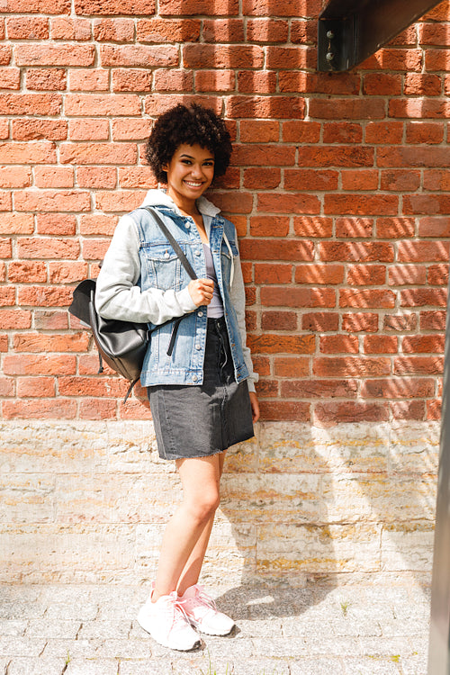 Portrait of a young smiling woman standing at brick wall, looking at camera