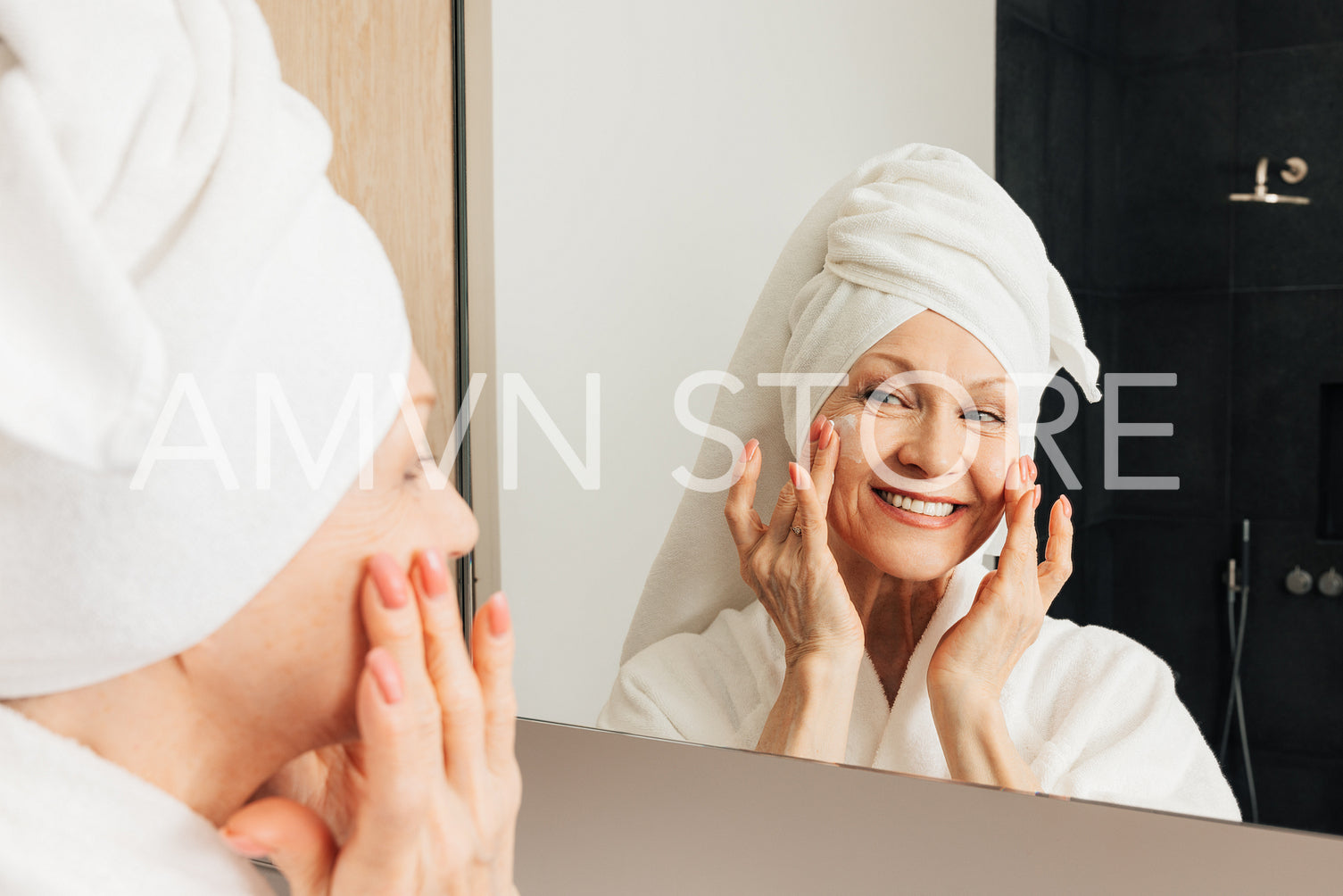 Smiling aged woman with a towel on a head applying facial cream on her cheeks in bathroom