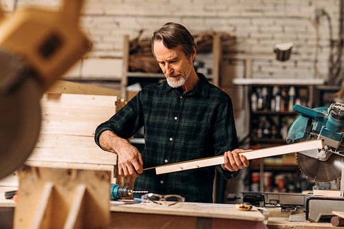 Male carpenter holding a wood plank and looking on measurement tape in a workshop