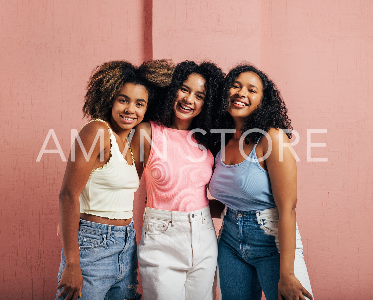 Three positive women with curly hair standing together against a pink wall and looking at the camera 