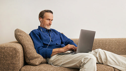 Senior man lying on a couch in the living room with a laptop on his hips