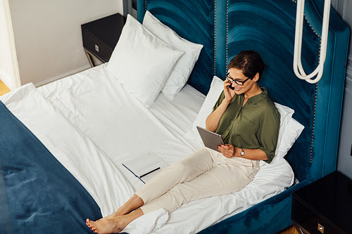 High angle view of woman in casual clothes lying on bed talking over mobile phone and looking on digital tablet