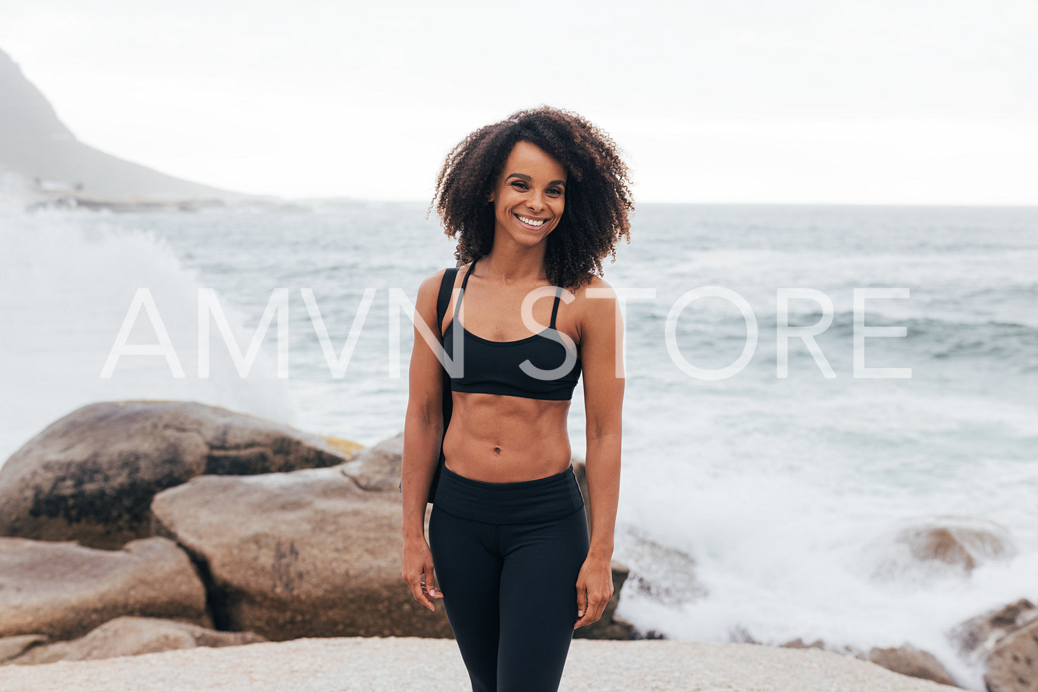 Smiling female relaxing after yoga. Muscular woman standing on rocks by ocean.