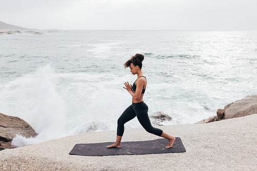 Side view of young female in fitness wear practicing yoga on mat by seaside