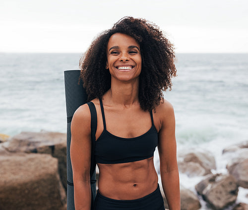 Muscular woman with yoga mat laughing while standing by ocean